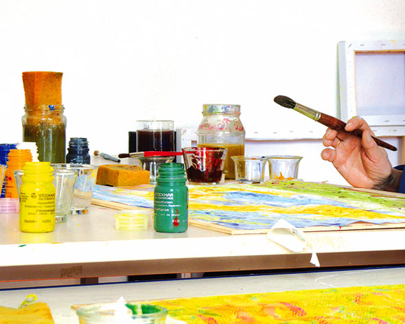 Eine Person sitzt an einem Arbeitsplatz mit Aquarellfarben und hält einen Pinsel in der Hand.